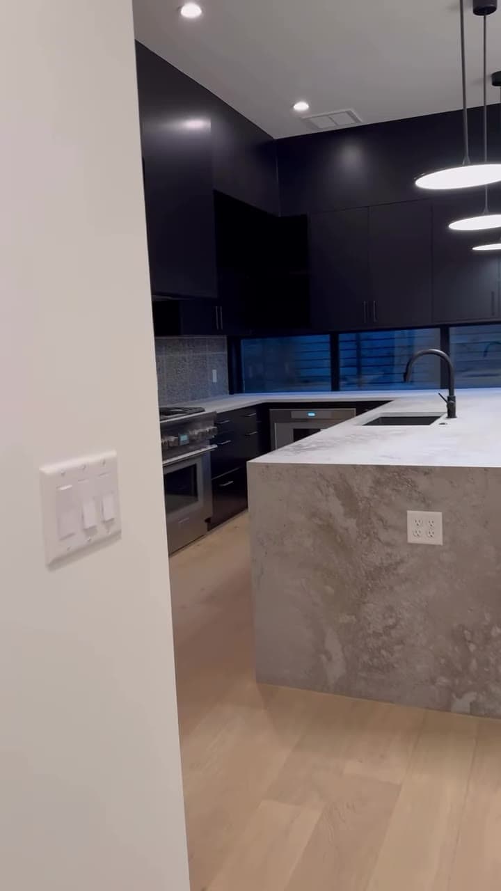Finished black kitchen viewed from the connected room with a waterfall island and pendant lighting